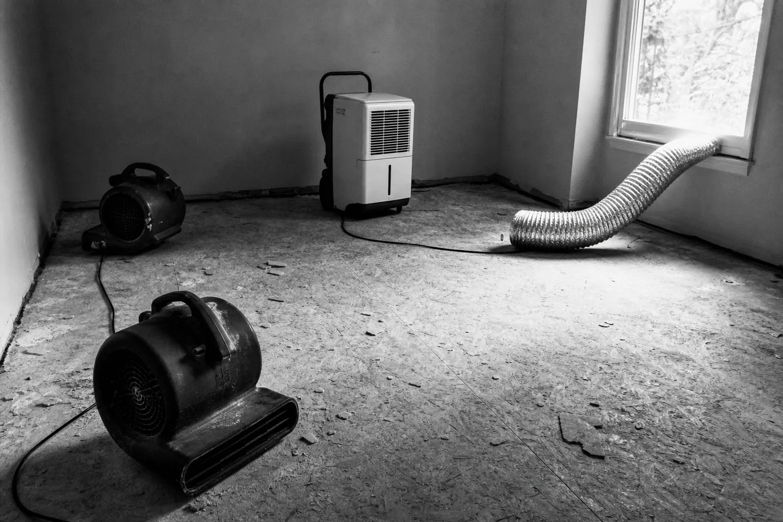 Two air movers and a dehumidifier running on a stripped subfloor in an empty room, with a length of ducting hose running to a window