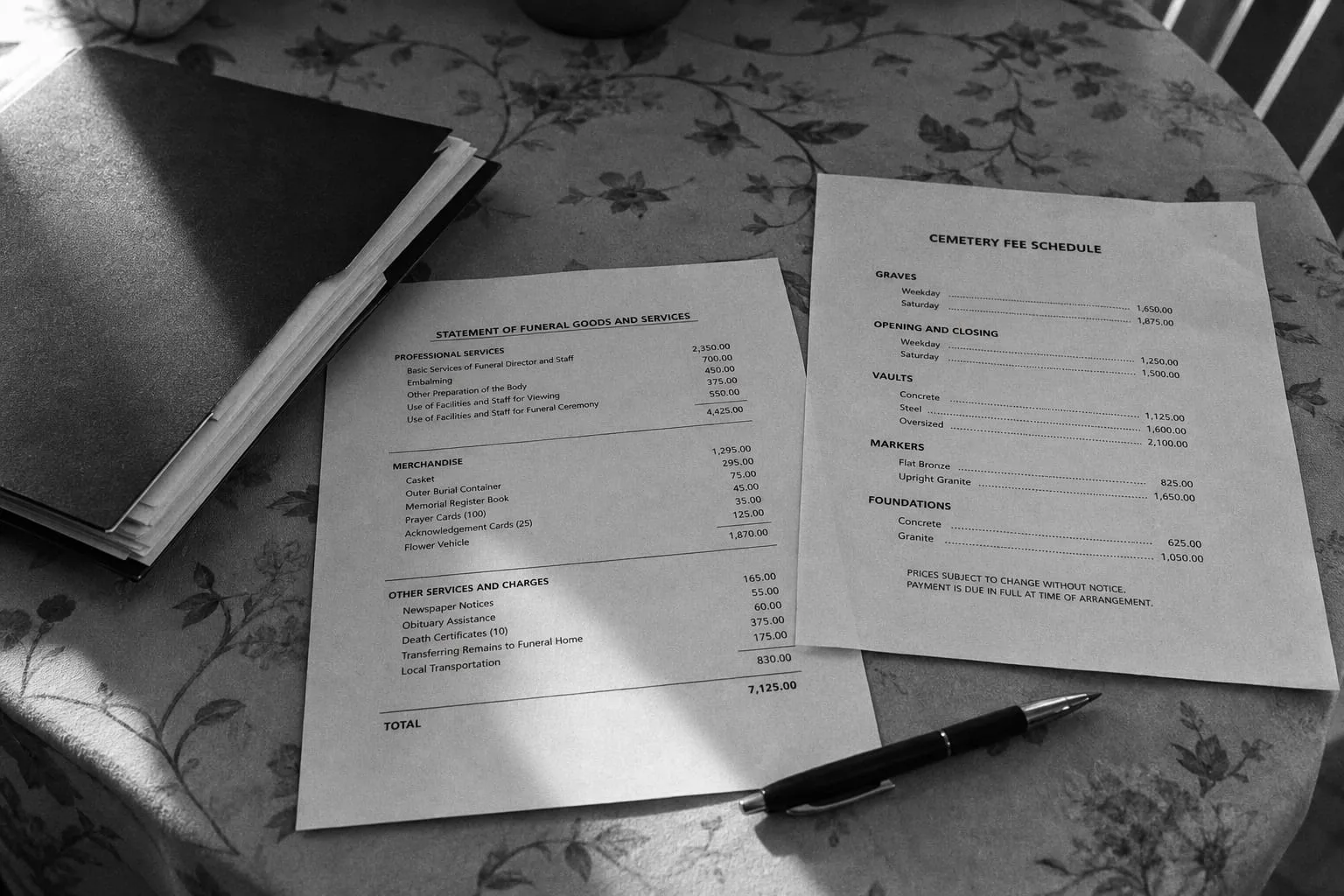 A printed itemized funeral goods and services statement and a separate cemetery fee schedule lying side by side on a kitchen table, with a pen and a folder o...