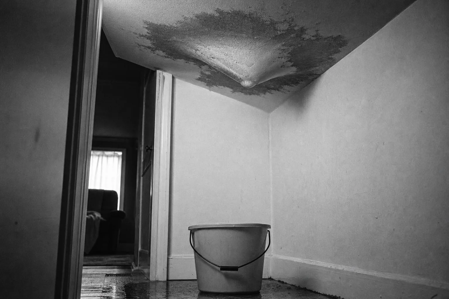 A ceiling with a sagging, water-stained patch of drywall bulging downward, a plastic bucket positioned on the floor beneath it
