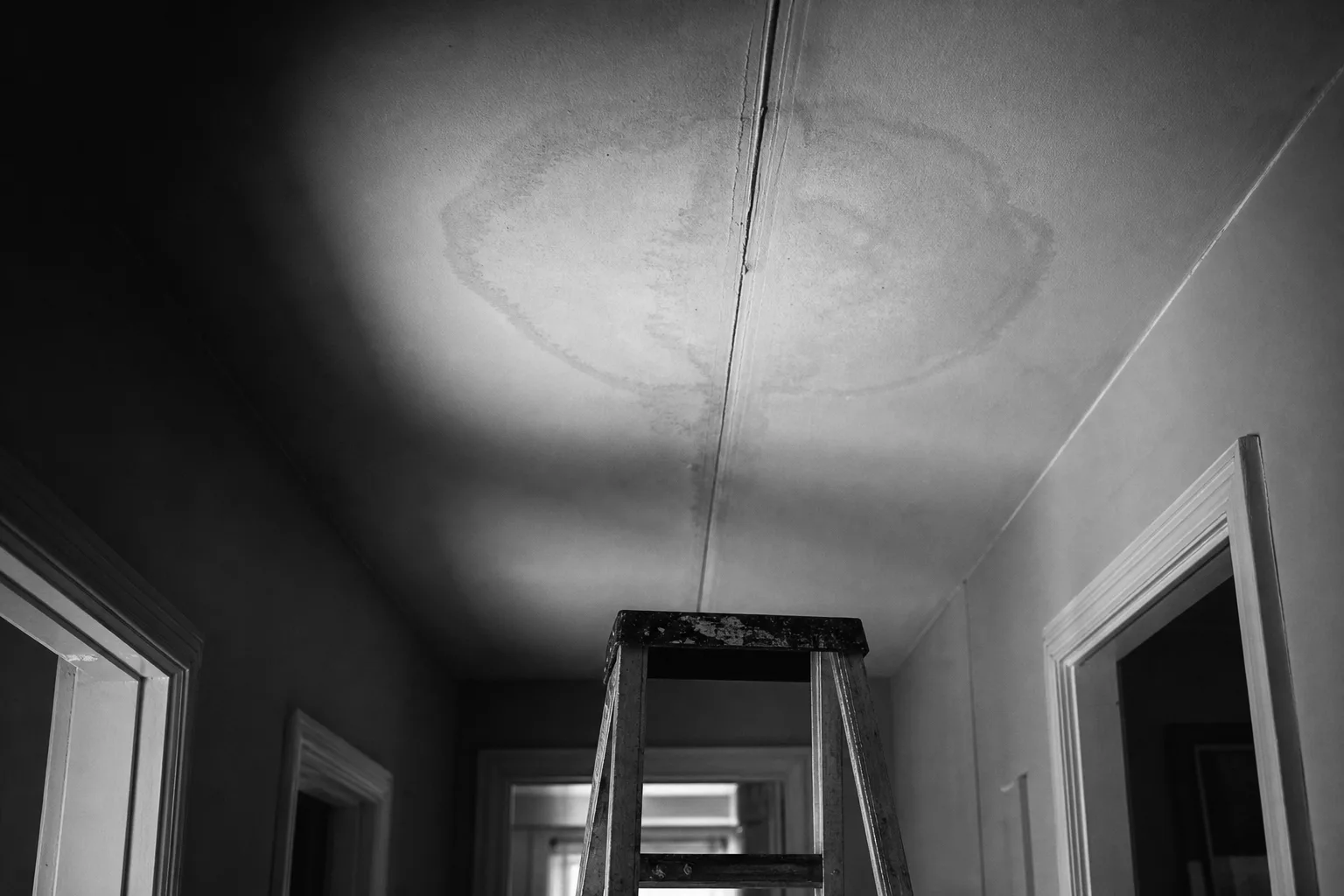 A hallway ceiling with a faint circular water stain spreading from a drywall seam, a stepladder standing beneath it