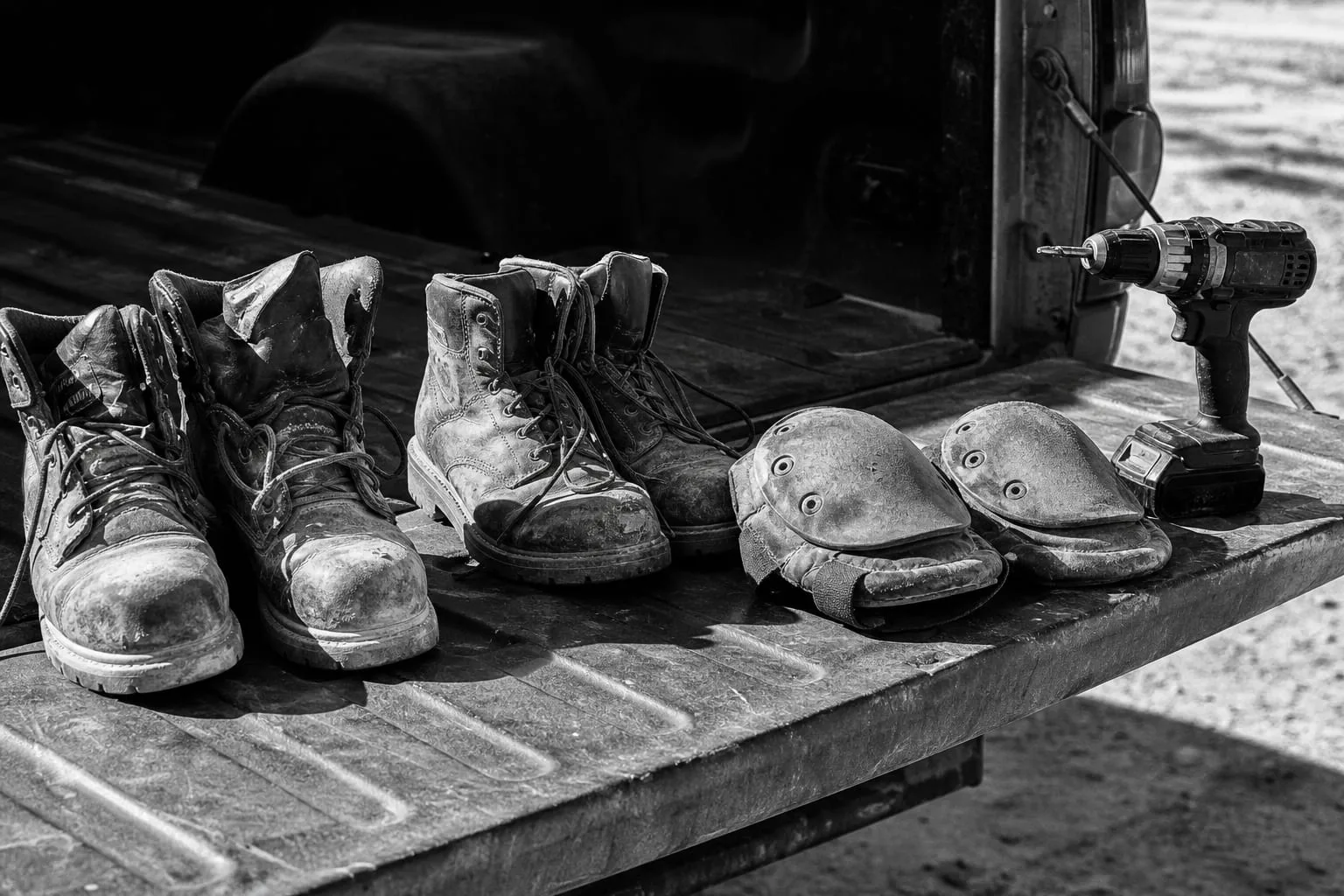Two pairs of worn work boots and a set of knee pads on the tailgate of a pickup beside a cordless drill