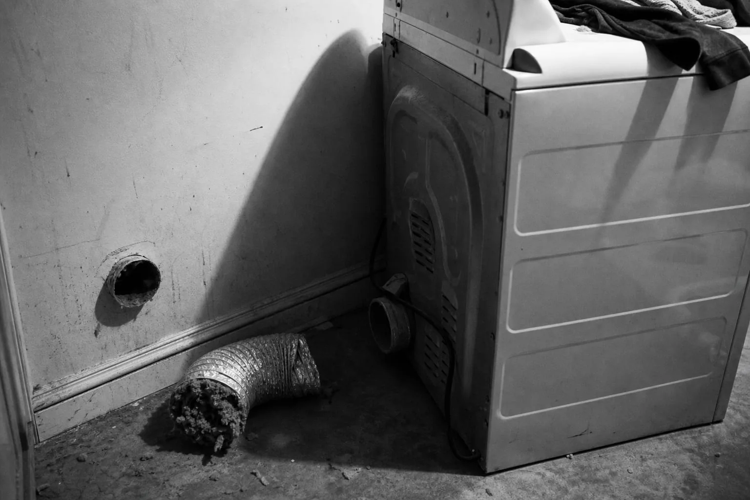 A clothes dryer pulled away from a laundry room wall with its flexible vent hose disconnected and packed with gray lint