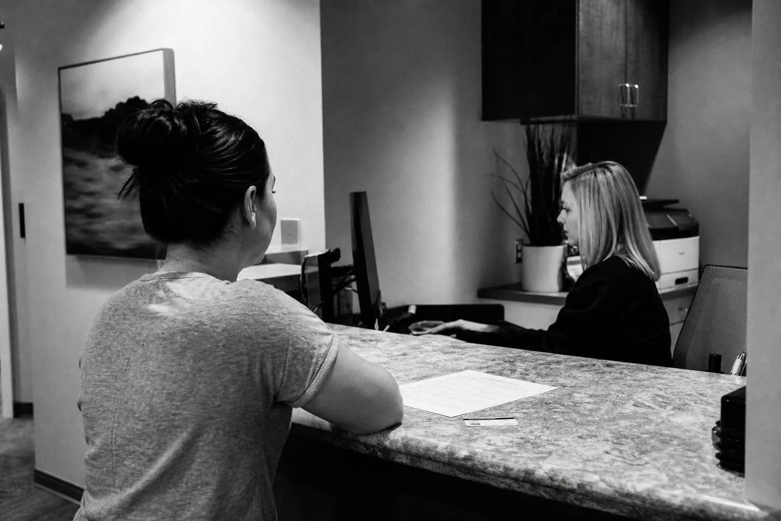 A patient seated at a private medical practice reception desk, insurance card and a paper intake form on the counter, receptionist working at a computer