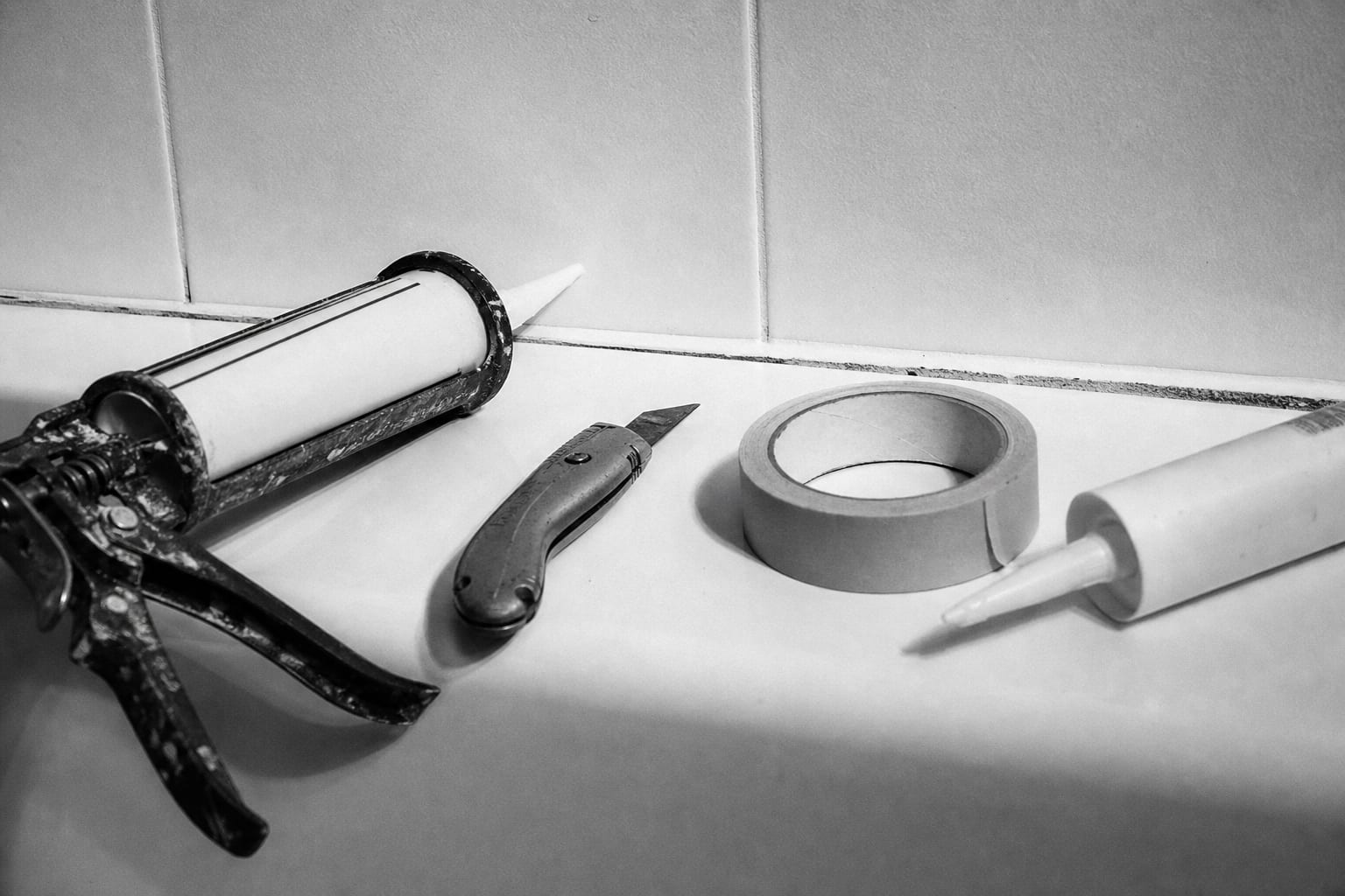 Close-up of the horizontal joint where a white bathtub meets tiled wall, with a caulk gun, utility knife, roll of painter's tape and a tube of silicone seala...