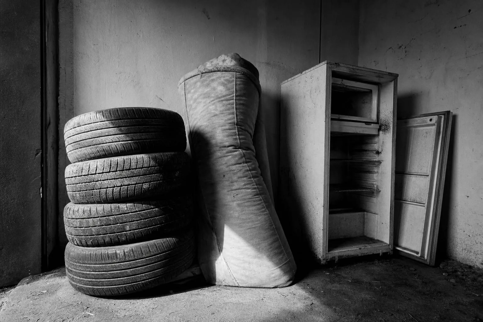 A garage corner stacked with four worn car tires, a rolled mattress leaning against the wall, and an old refrigerator with its door removed