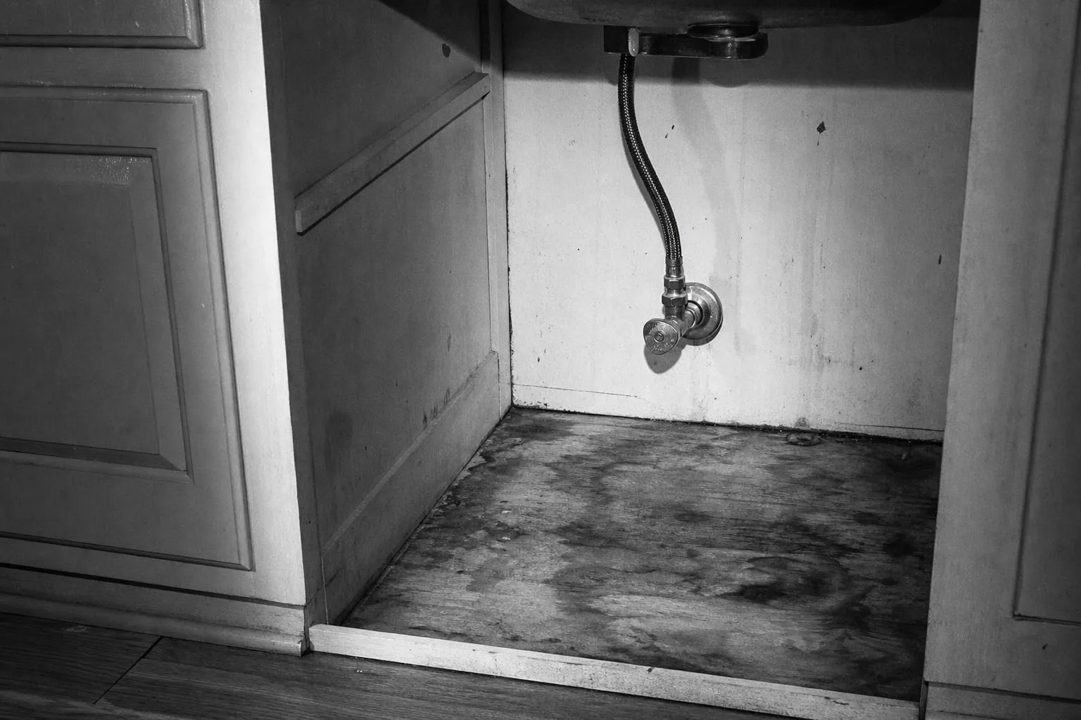 The kickspace beneath a kitchen sink opened up, showing a stained subfloor, a braided supply line and a shutoff valve