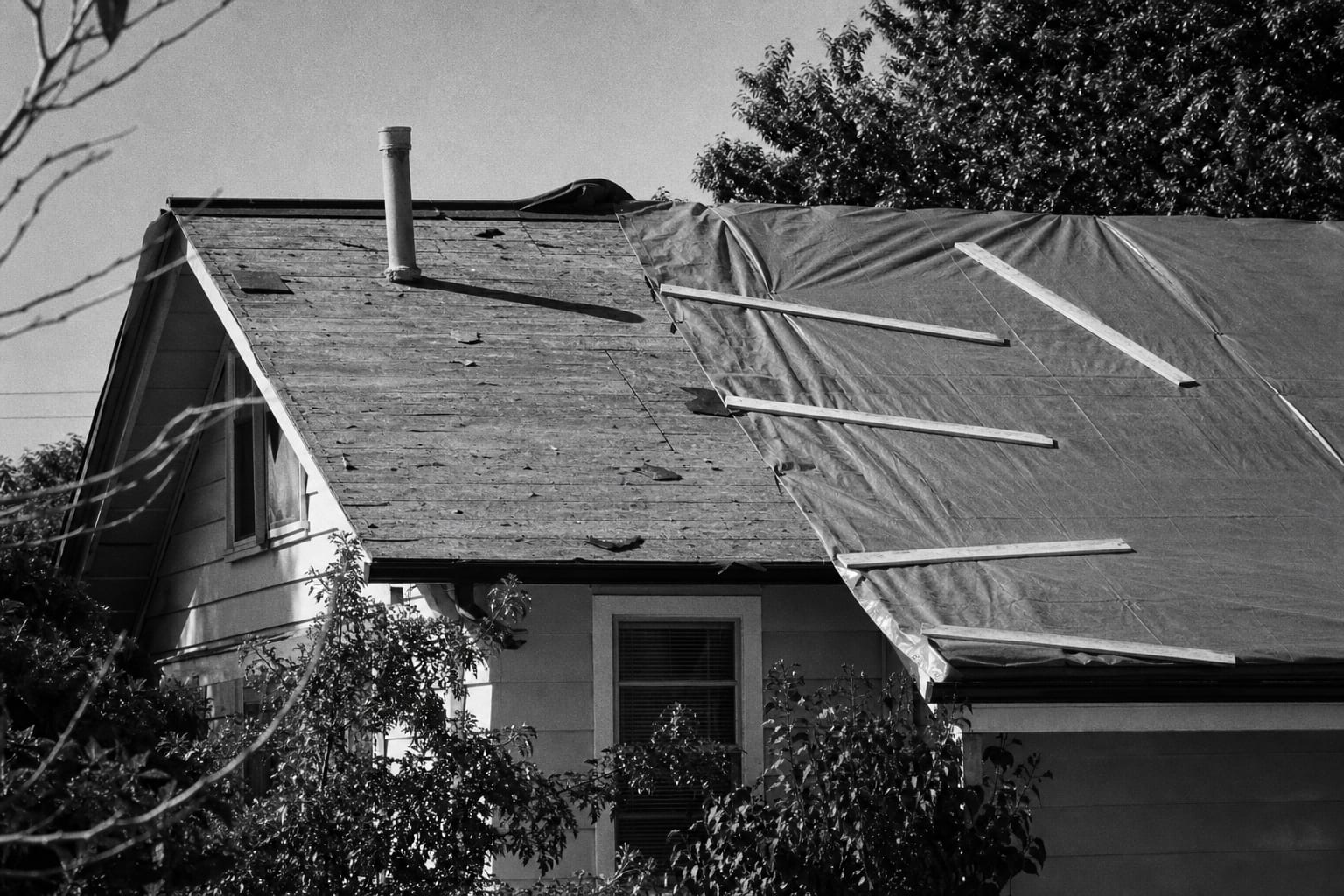 A residential roof partly stripped to bare underlayment with a blue tarp weighted down by lumber across one slope