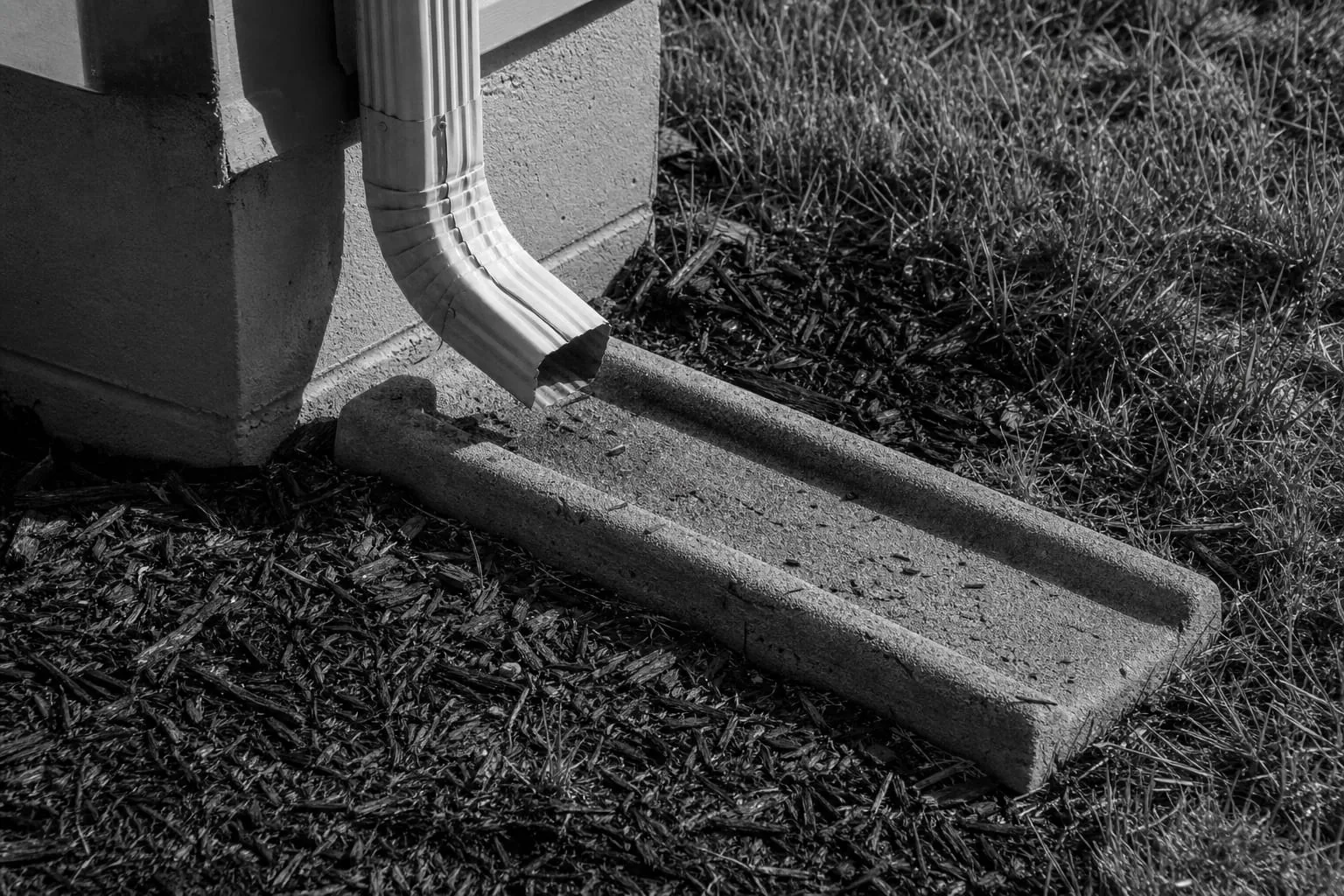 A downspout elbow discharging onto a concrete splash block at the corner of a house foundation, with mulch and grass alongside