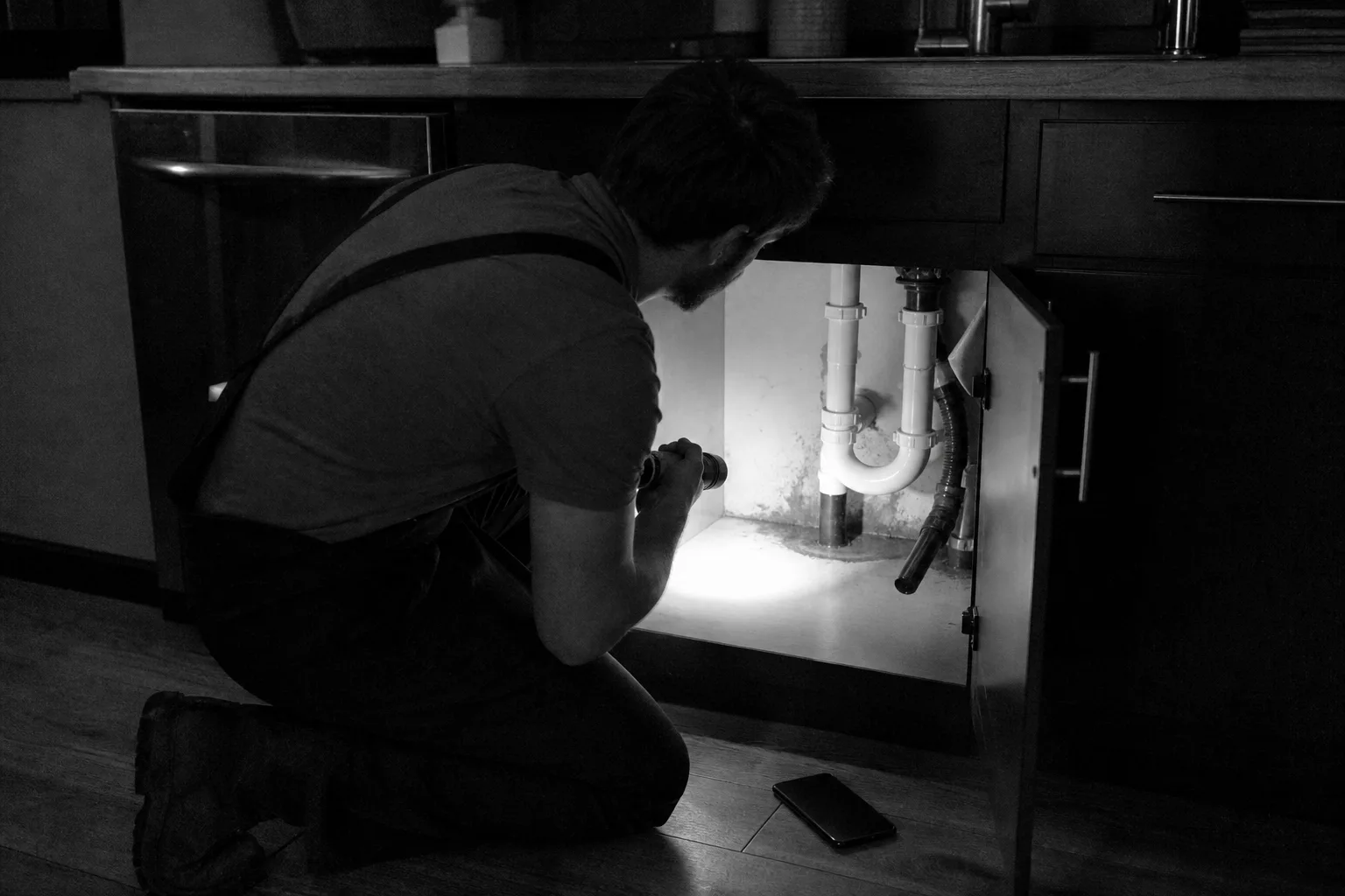 A cleaner kneeling beside an open kitchen sink cabinet, flashlight in hand, examining the base of the cabinet where the pipe enters, with a phone resting on...